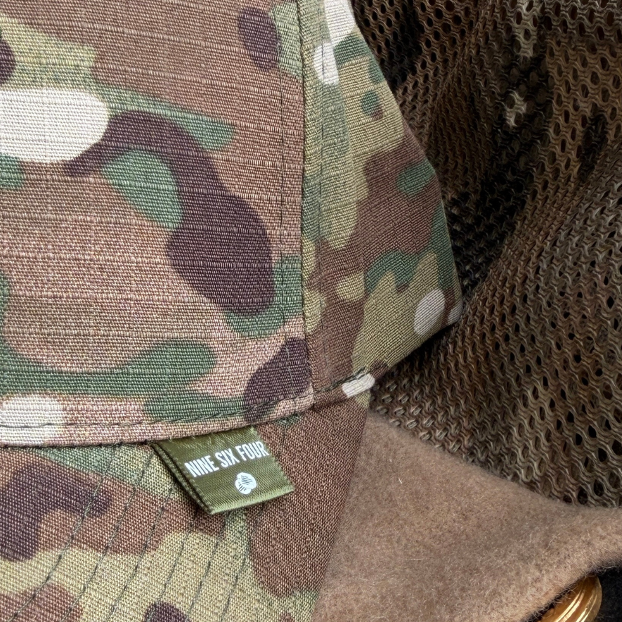 Close-up of a camouflage baseball cap with a small green 'NINE SIX FOUR' brand tag on the brim, layered over a tan fleece and CAMO mesh.