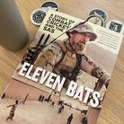 A book cover featuring a man in military fatigues with a hat on, holding a bat, with the title 'Eleven Bats' prominently displayed. The background includes a mug and several coins on a wooden surface.