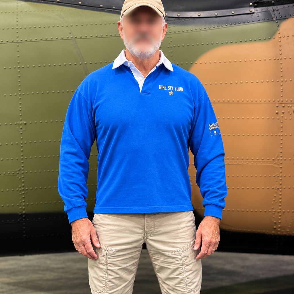 A man wearing a bright blue long-sleeve polo shirt with 'NINE SIX FOUR' printed on the chest, khaki cargo pants, tan work boots, and a tan baseball cap, standing in front of an aircraft nose.