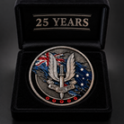 A commemorative military coin displayed inside an open black presentation box. The coin features a silver-toned winged dagger emblem with a banner reading 'WHO DARES WINS,' blue and red flag elements, and small red poppy details around the bottom edge.